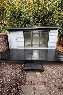 A small, light gray luxury bespoke garden room with a black deck and sliding glass doors, situated between two wooden fences and surrounded by trees in Cambridgeshire.