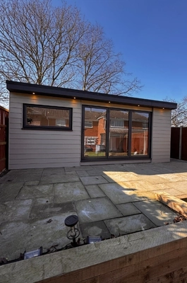 A modern, luxury bespoke garden room in Hertfordshire with large sliding glass doors and horizontal cladding, set on a stone patio with a wooden border, under a clear blue sky.