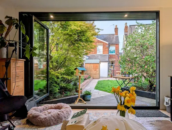 A room with large open sliding doors leads out from a luxury bespoke garden room in Hertfordshire, overlooking trees and a path, with a dog bed, flowers, and books in the foreground.