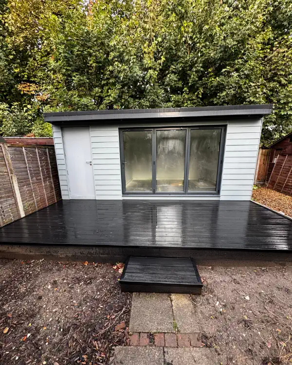 A small, light gray luxury bespoke garden room with a black deck and sliding glass doors, situated between two wooden fences and surrounded by trees in Cambridgeshire.