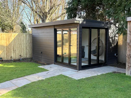 Luxury bespoke garden rooms with glass doors and a chair inside, situated on a lawn with a stone path leading to the entrance, surrounded by a wooden fence and trees.