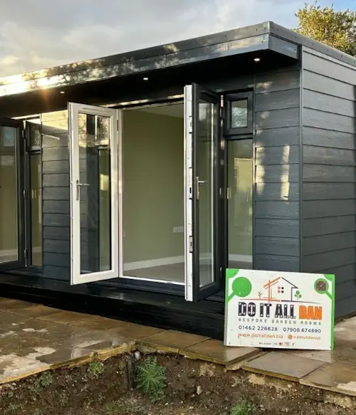 A modern garden room with open double glass doors, dark exterior cladding, and a "Do It All Dan" sign in front, showcasing luxury bespoke garden rooms in Hertfordshire.
